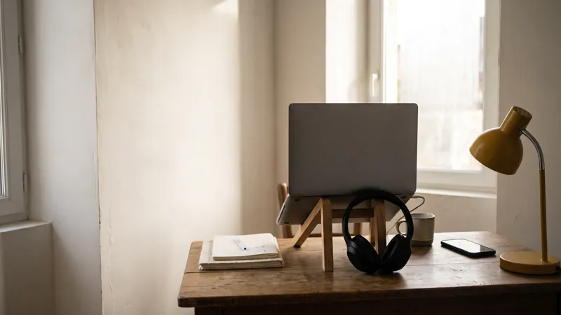 Coin bureau près d'une fenêtre avec un ordinateur portable sur support, un casque audio, un smartphone et une lampe de bureau jaune